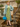 Model wearing a sage green coat and yellow floral dress holding the marine teal blue frame handbag beside stone steps